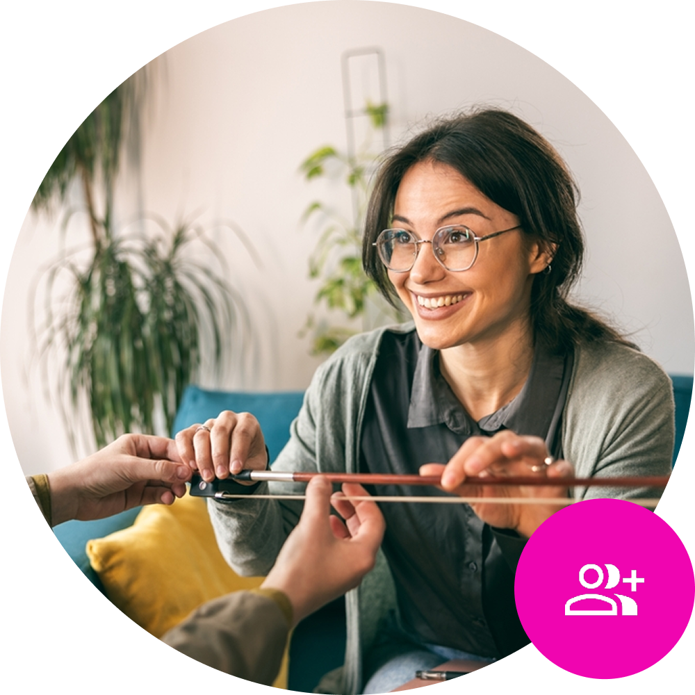 Female music teacher holding violin bow and looking at student across the table - CRM Icon in the bottom right of the screen on brand pink background