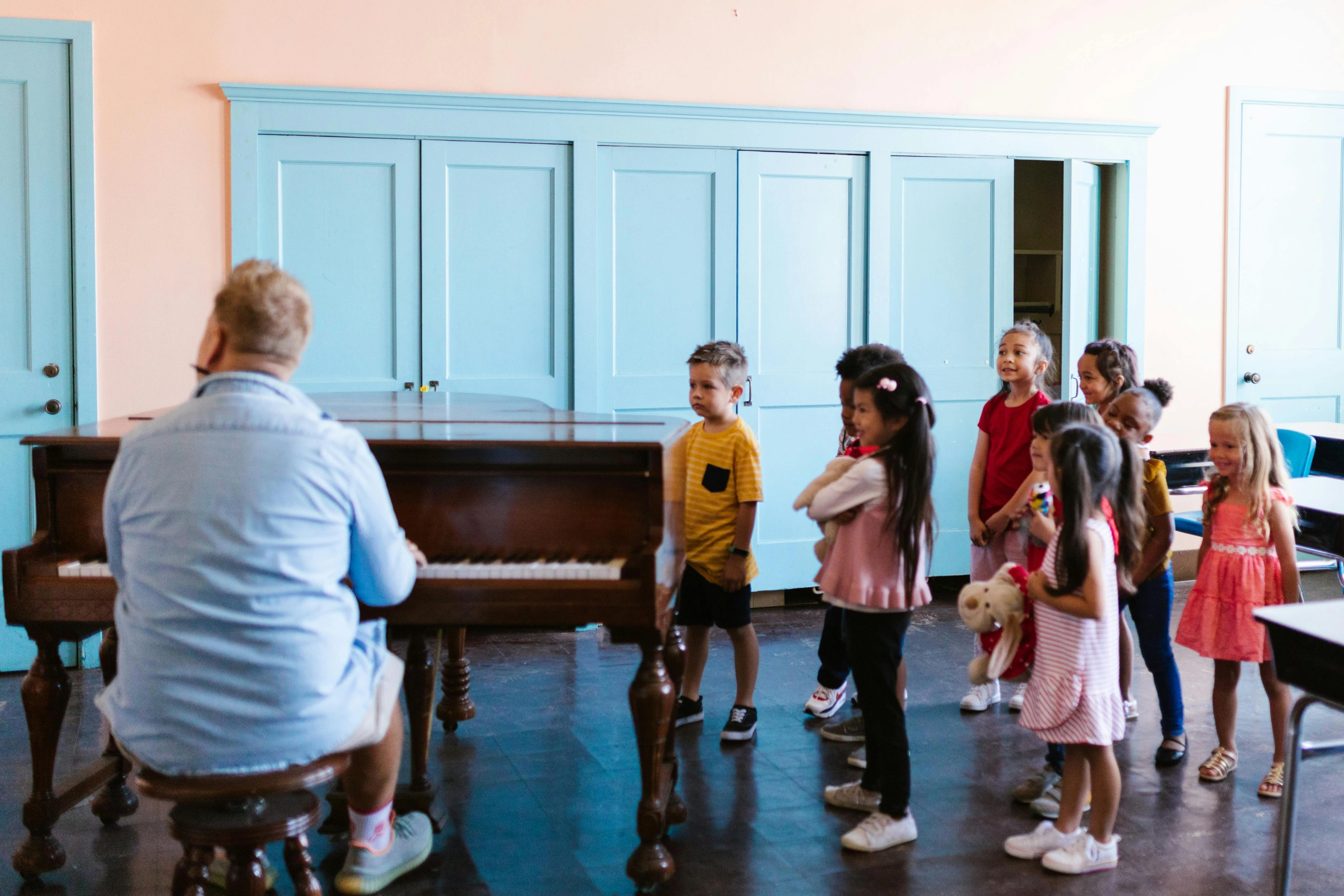 A group of young children gathers around a piano as their teacher leads a fun classroom music session.