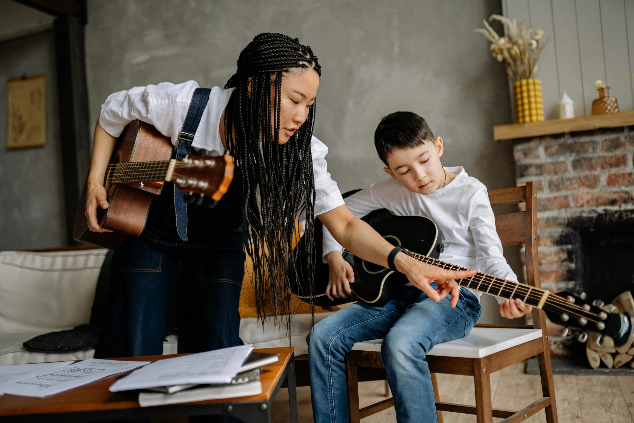 A guitar teacher guides a young student through finger placement during a lesson at home.