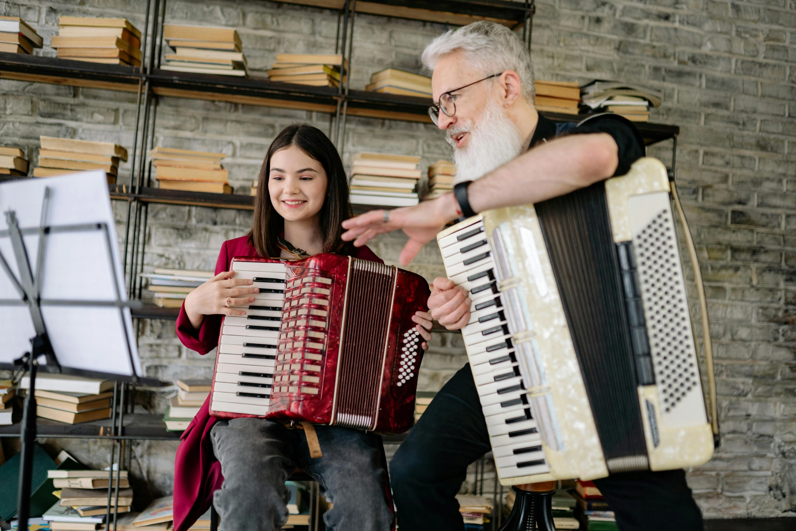 A bearded music teacher gives accordion instruction to a smiling teenage student during a lesson.