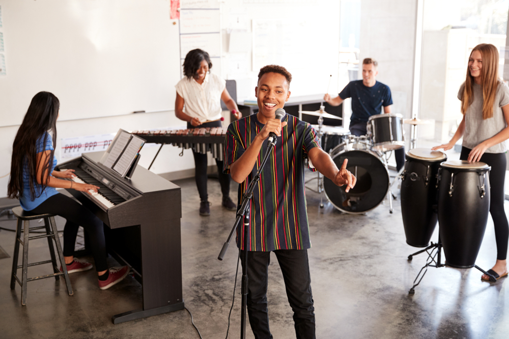A group of diverse students rehearses in a bright, modern music classroom / performing arts school, performing vocals, piano, drums, and percussion.