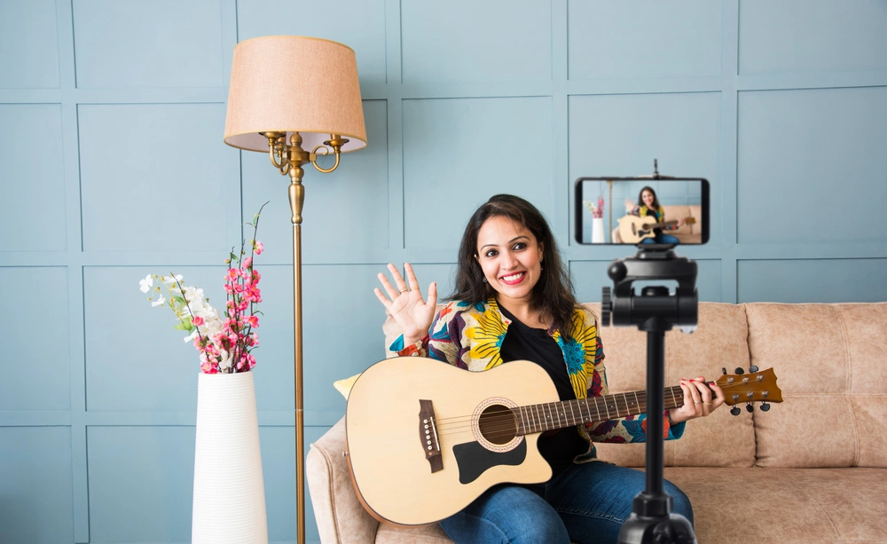 A cheerful guitar instructor records an online lesson or livestream from home, engaging directly with her virtual students.