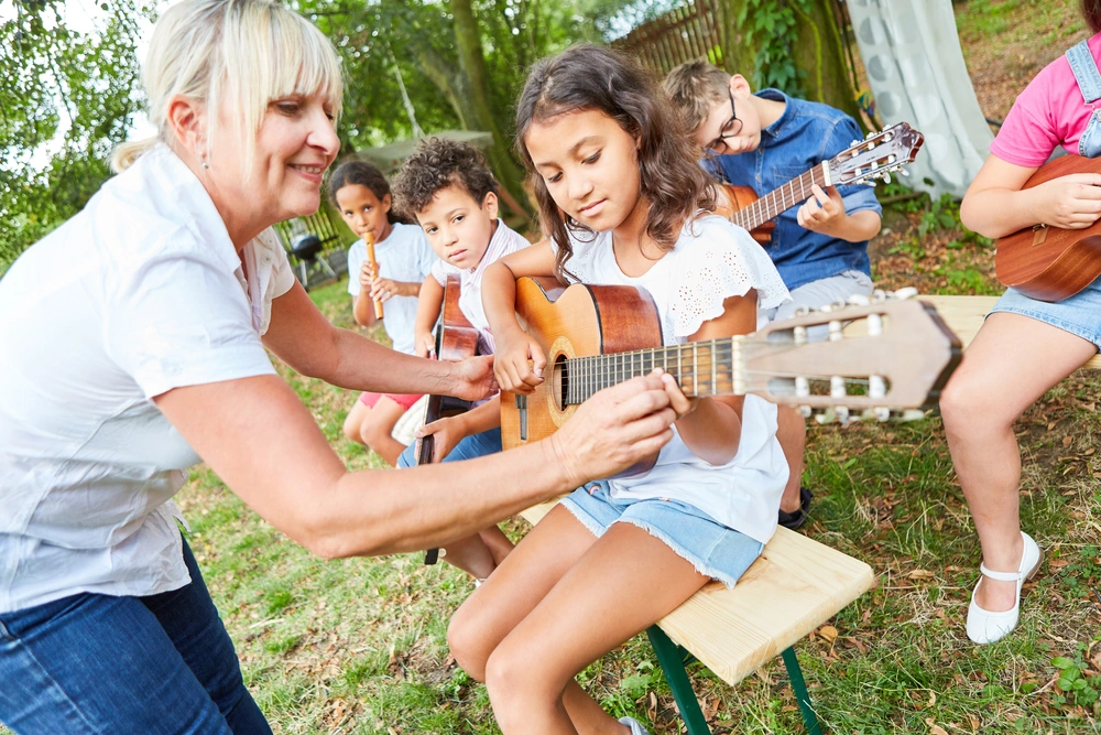 A group of young students participates in an outdoor guitar lesson, guided by an encouraging instructor.