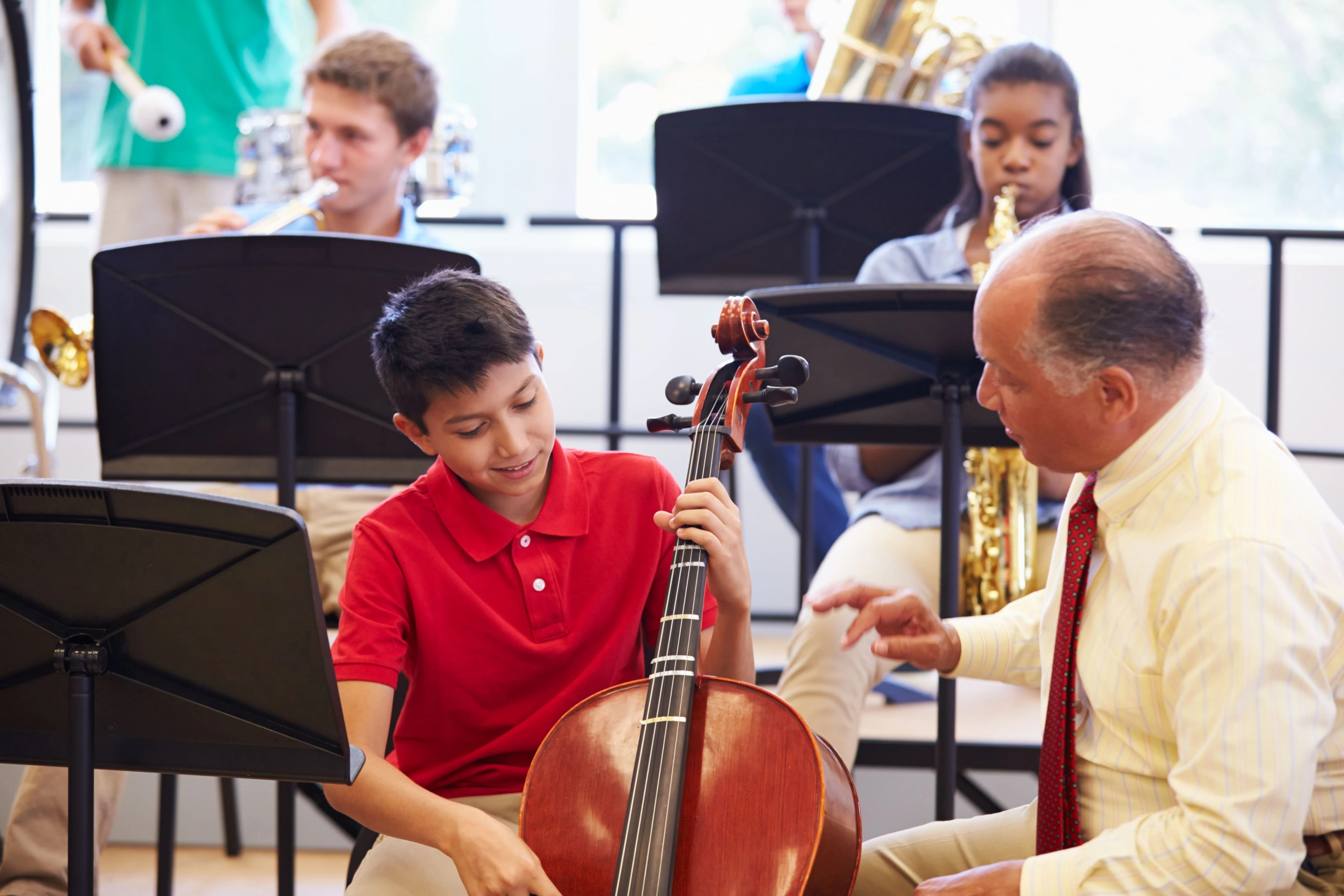 A music teacher provides instruction to a student holding a cello during a band class rehearsal.