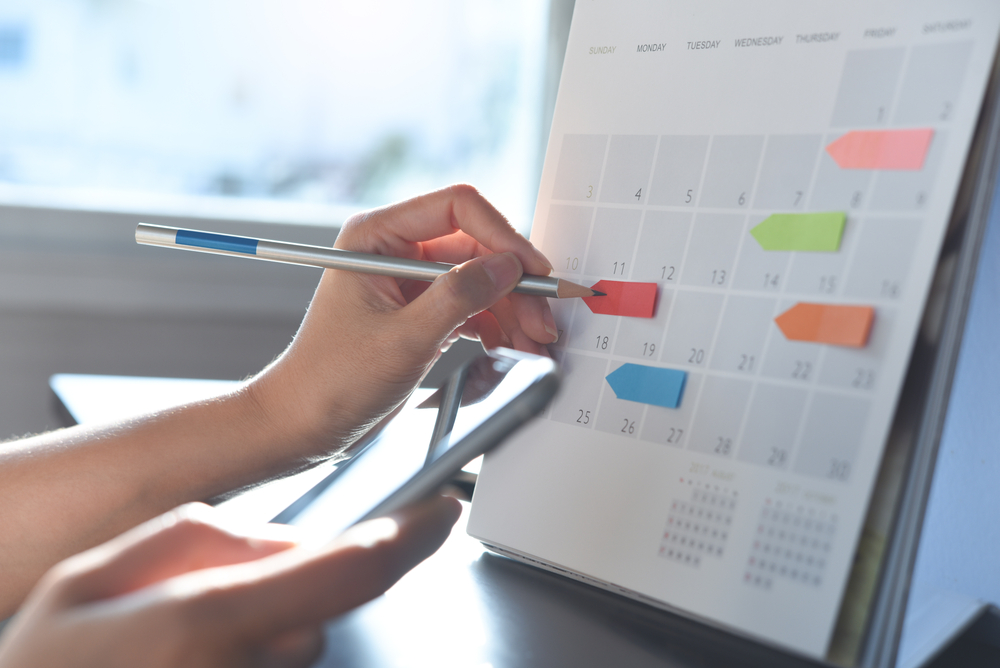 A person updates a color-coded calendar while referencing their phone, organizing upcoming events and schedules.