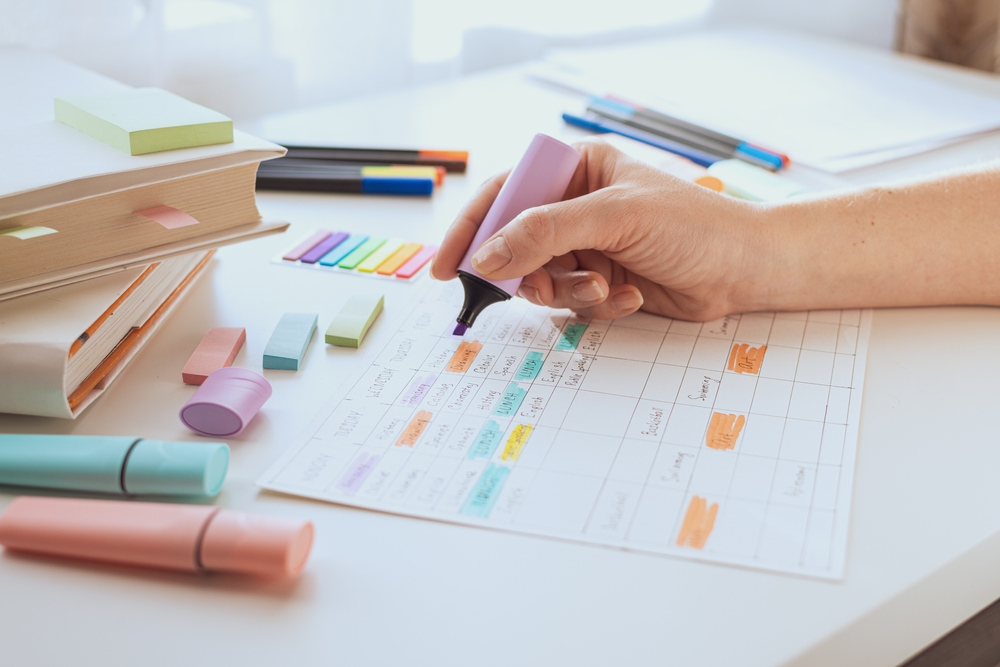 A hand highlights a colorful weekly schedule with markers, surrounded by planning tools and study materials.