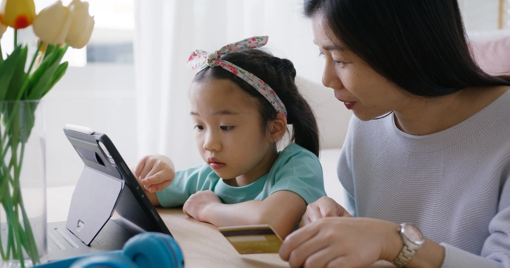A parent and child sit together at a tablet, navigating an online payment or enrollment form with a credit card in hand.