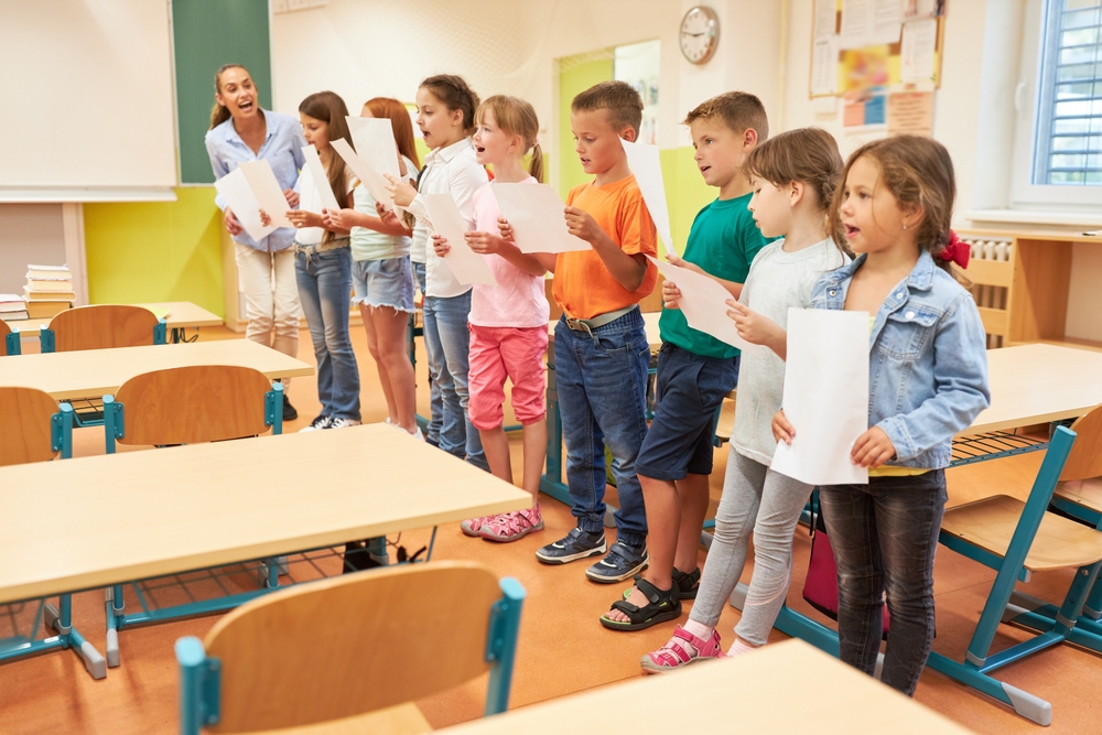 A group of elementary students practices singing in a classroom, holding lyric sheets and following their enthusiastic teacher.
