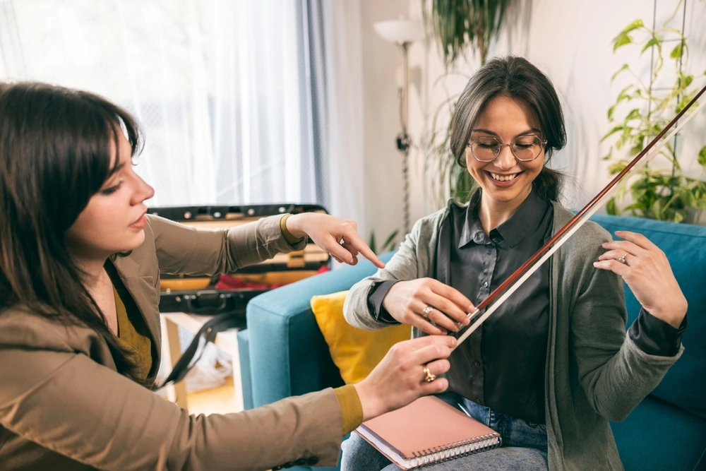 An instructor and student share a hands-on violin lesson in a relaxed home environment, focusing on bow technique.