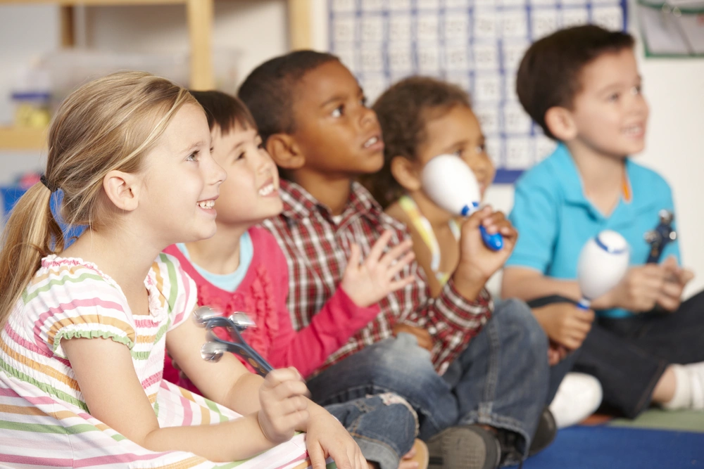 A group of smiling young children plays handheld percussion instruments during an early childhood music class.