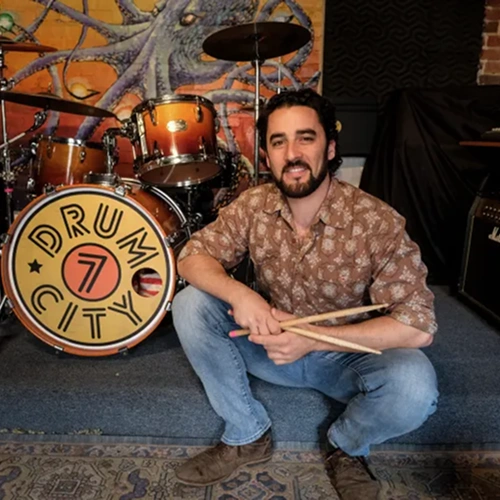 Seven Drum City owner sitting in front of drums holding drum sticks