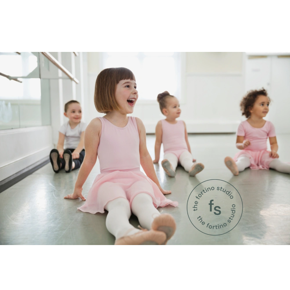 A group of young children sit on the floor of a bright dance studio, smiling and relaxing during a ballet class. In the foreground, a girl wearing a pink leotard and skirt with white tights sits with her legs stretched out, laughing and looking to the side. Other children in similar dance attire sit behind her, also smiling. A mirrored wall and ballet barre are visible along the side, and sunlight fills the room. The Fortino Studio logo is in the corner.